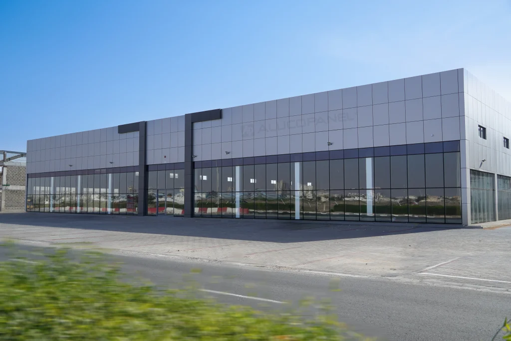 Angled view of commercial warehouse building clad in ALUCOPANEL® light grey composite panels with full-height reflective glass curtain wall in UAE