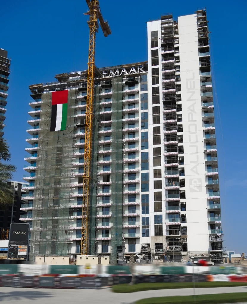 Street-level view of an Emaar development under construction in Dubai showing balconies clad in Alucopanel panels and a UAE flag displayed on scaffolding