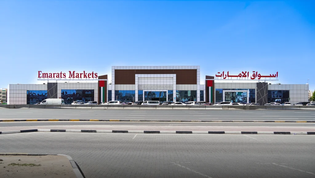 Front elevation of Emarats Markets retail complex displaying symmetrical white and brown Alucopanel facade, dual English and Arabic rooftop signage, UAE flags, full parking area, and a clear blue sky backdrop